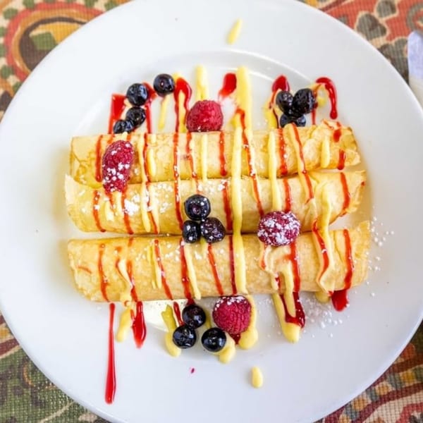 Three crepes drizzled with yellow and red sauces, topped with raspberries and blueberries on a white plate.