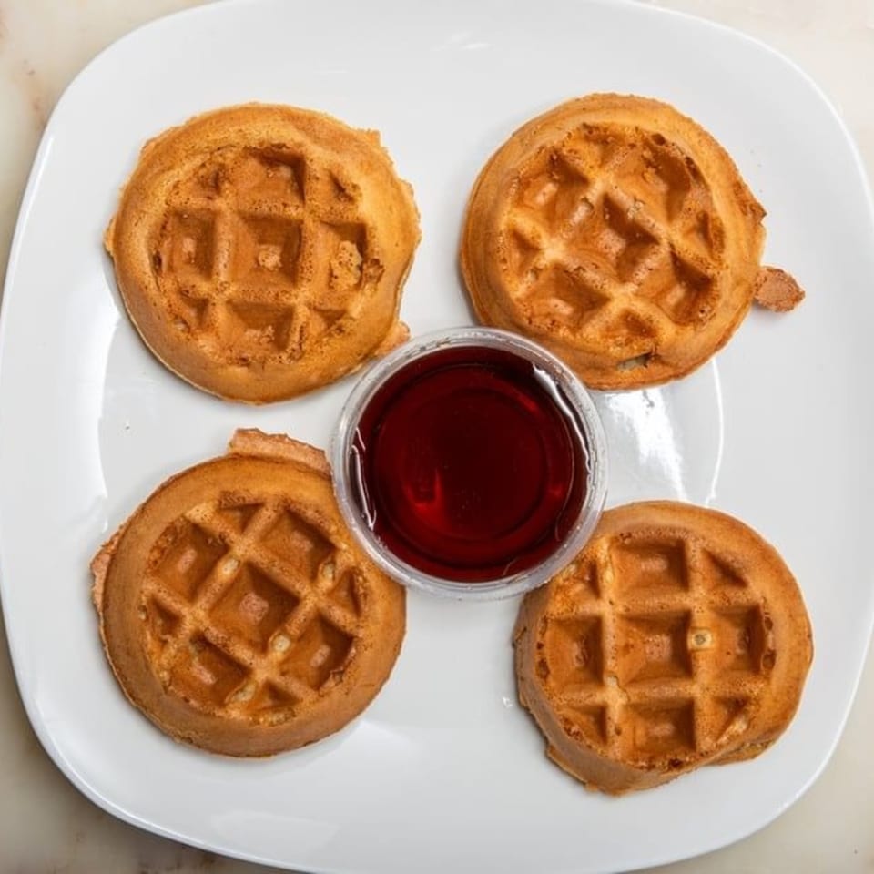 Four golden-brown mini waffles surround a small cup of maple syrup on a white plate.
