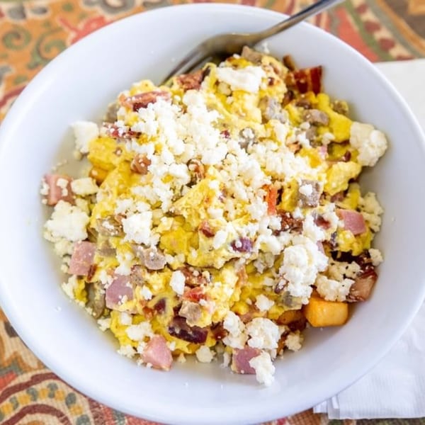 A white bowl of scrambled eggs with bacon, ham, sausage, and crumbled feta cheese.