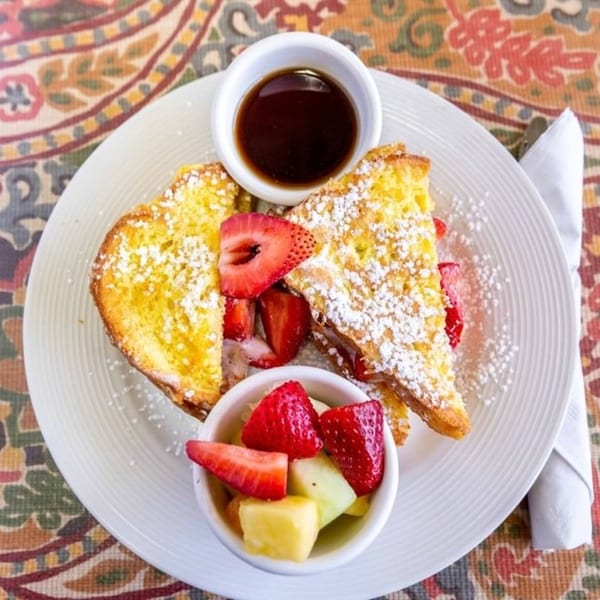 Golden French toast with powdered sugar, fresh strawberries, fruit cup, and syrup on a white plate.